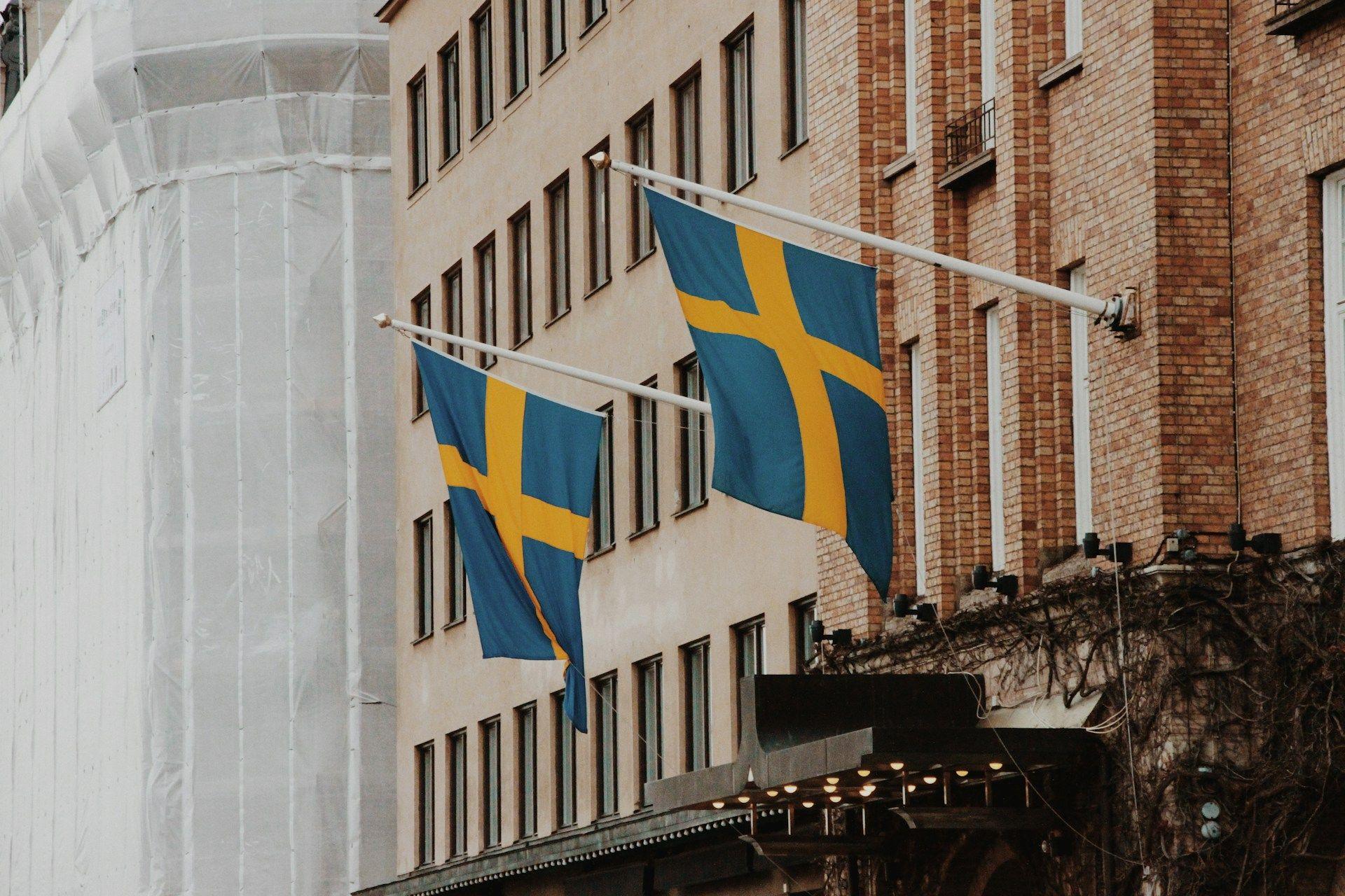 Two blue and yellow Swedish flags wave proudly from a traditional brick building facade.