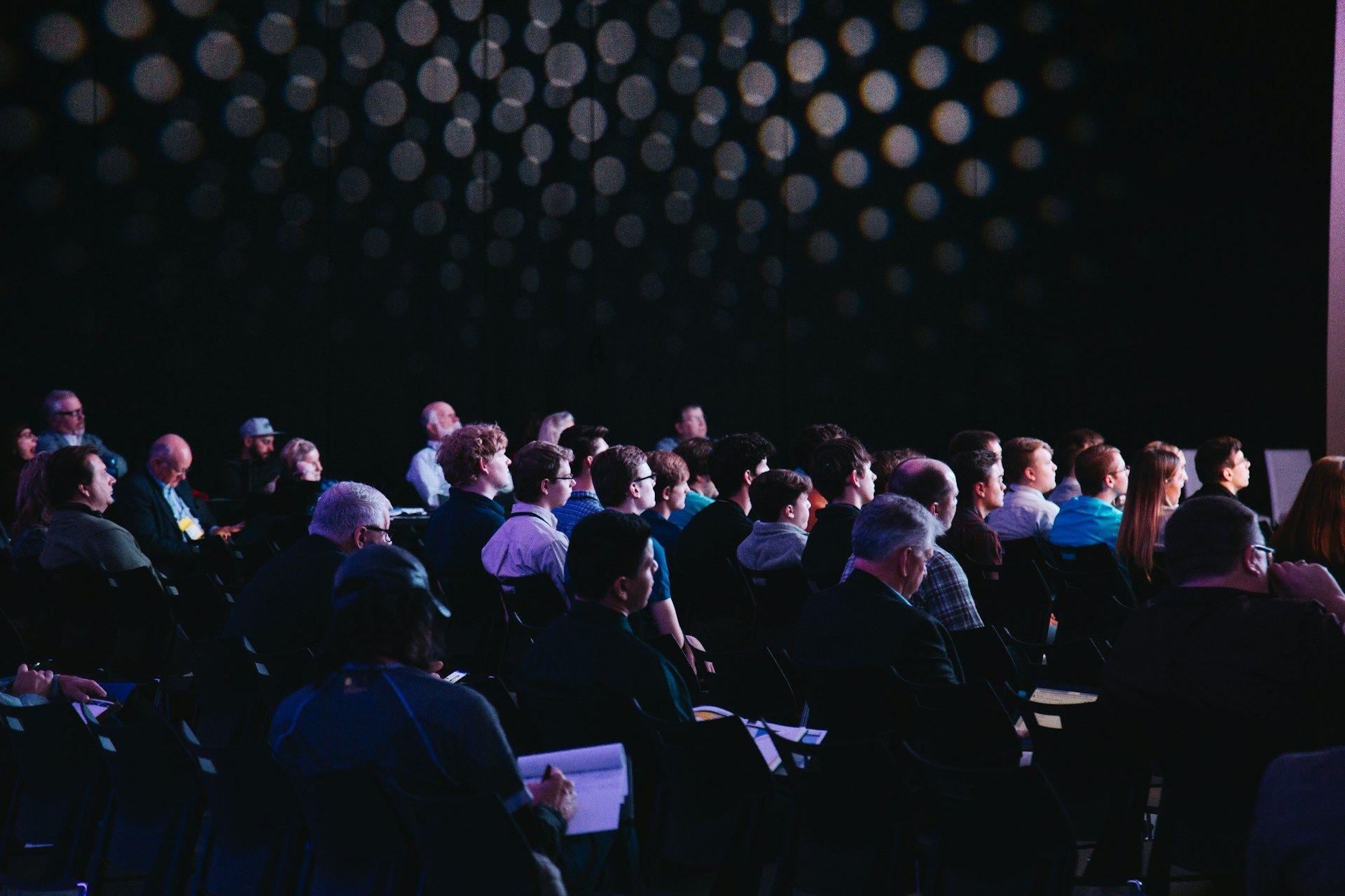 Audience attending a dark conference room with a patterned wall.